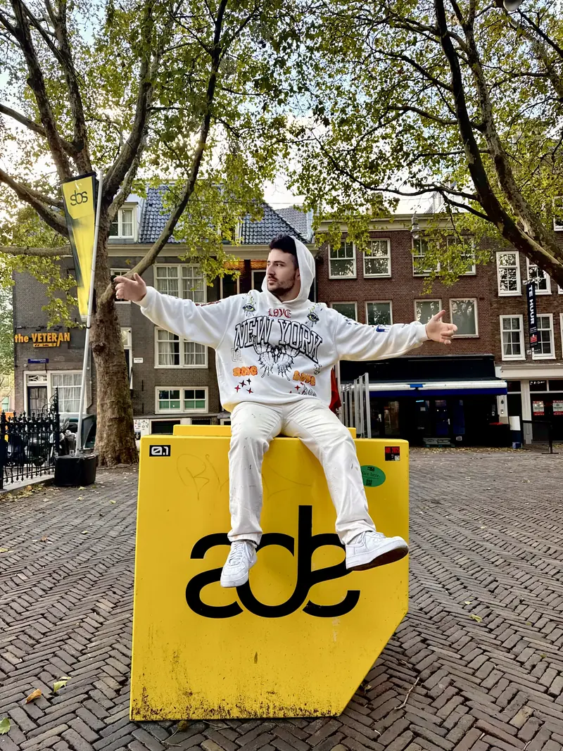 Yohann Warren sitting arms wide on the yellow ADE logo block on a square in Amsterdam