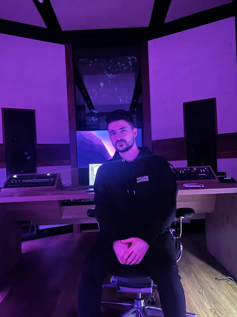 Yohann Warren seated in a purple-lit music studio in front of the mixing desk, wearing a Yohann Warren hoodie